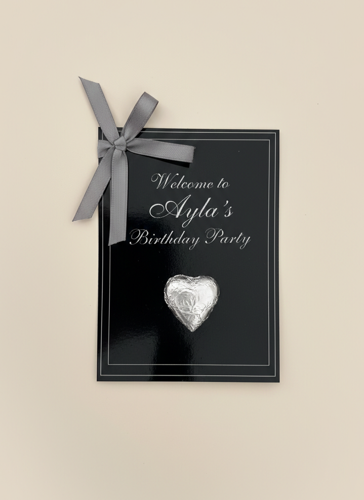 Black birthday party invitation with silver heart and bow on a beige background