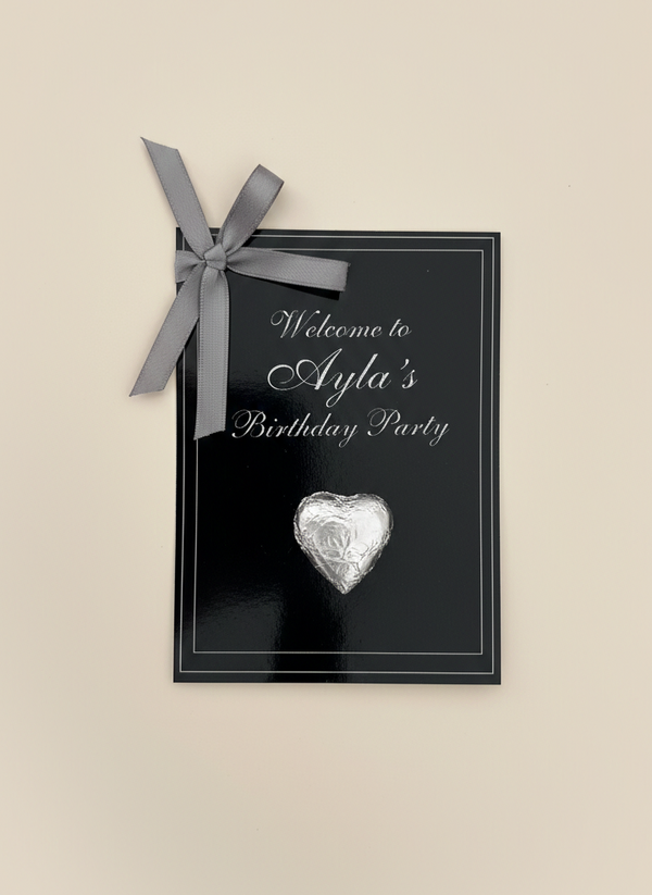 Black birthday party invitation with silver heart and bow on a beige background