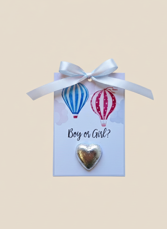Heart-shaped gold charm on a card with hot air balloons and 'Boy or Girl?' text, tied with a white ribbon.