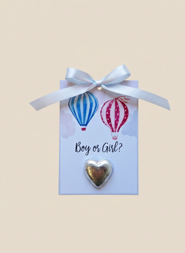 Heart-shaped gold charm on a card with hot air balloons and 'Boy or Girl?' text, tied with a white ribbon.