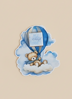 Sticker of a teddy bear in a hot air balloon with a blue ribbon on a beige background