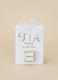 White gift bag with gold text and a small square card on a beige background