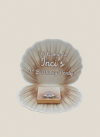 Seashell-shaped party favor with 'Welcome to Inci's 1st Birthday Party' text on a beige background
