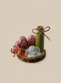 Decorative arrangement with a green candle, pink eggs, and a white figurine on a wooden base against a beige background.