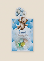 Party favor with teddy bear and balloons on a blue background, featuring the text 'Jamal is turning One'.