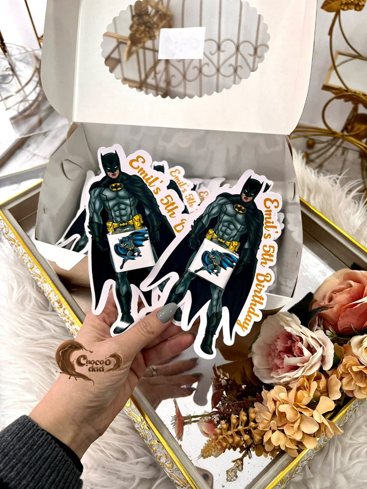 Two Batman-themed birthday stickers held by a hand with a tattoo, placed on a decorative box with flowers.