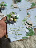 Hand holding a small card with a design of eggs and a person wearing a hat, on a blurred background.