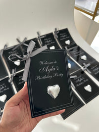 Black invitation card with silver ribbon and decorative stone held by a hand, with more cards in the background.