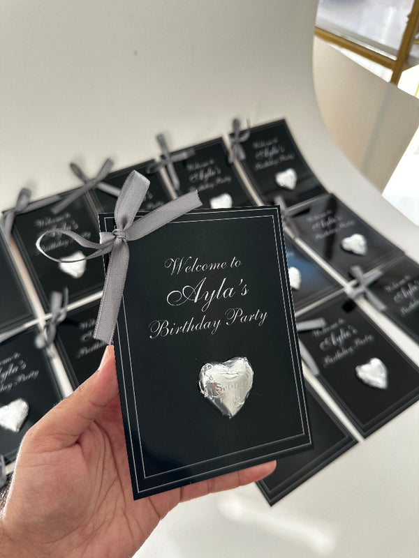 Black invitation card with silver ribbon and decorative stone held by a hand, with more cards in the background.