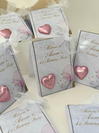 Party favor boxes with pink heart-shaped decorations and personalized text on a white surface.