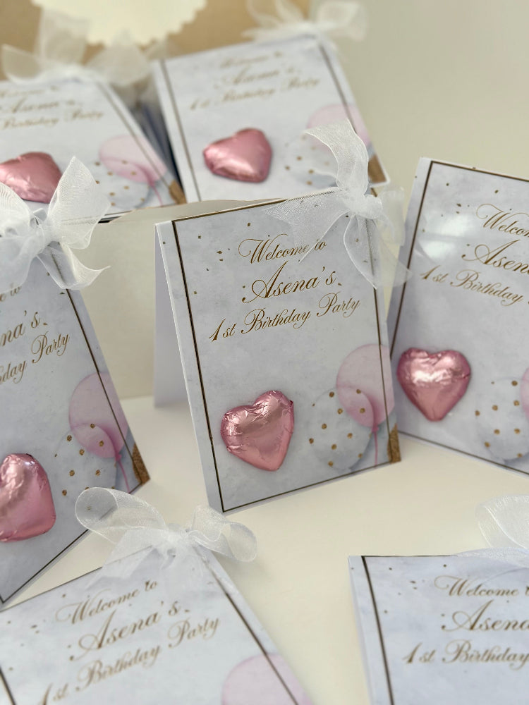 Party favor boxes with pink heart-shaped decorations and personalized text on a white surface.