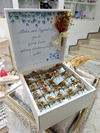 Decorative box with dried flowers and a message inside, set against a blurred indoor background.