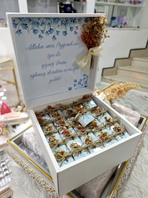 Decorative box with dried flowers and a message inside, set against a blurred indoor background.