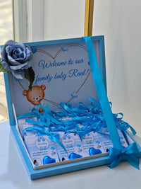 Gift box with baby items and blue ribbons, featuring a teddy bear and text 'Welcome to our family baby Raul'.