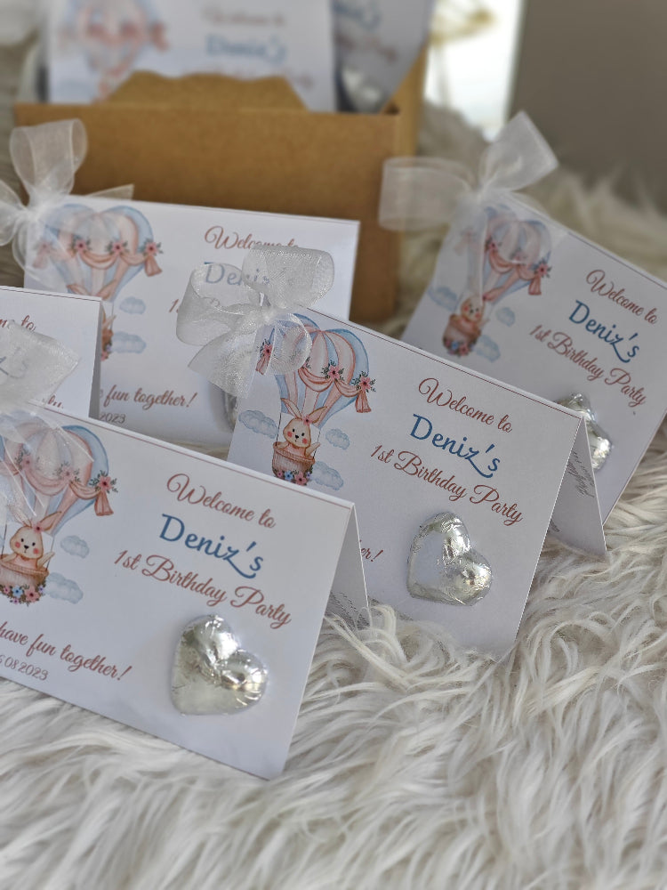 Party favors with heart-shaped decorations and 'Welcome to Deniz's 1st Birthday Party' text on a fluffy white surface.