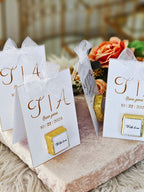 White cards with gold text and ribbons on a decorative surface with flowers in the background