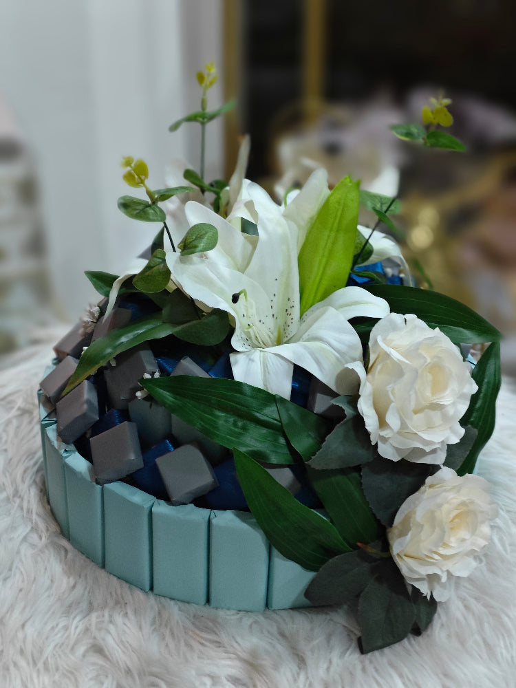 Decorative flower arrangement with white flowers and green leaves in a blue container on a soft surface.
