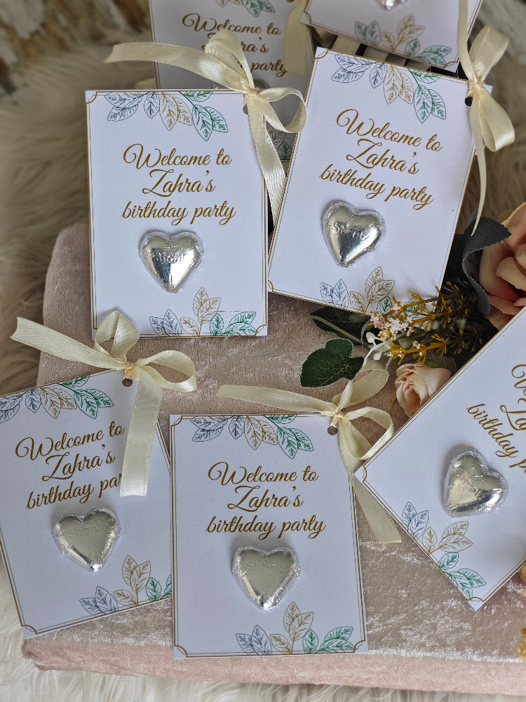 Silver heart-shaped earrings on cards with 'Welcome to Zahra's birthday party' text, arranged on a textured surface.