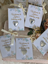 Silver heart-shaped earrings on cards with 'Welcome to Zahra's birthday party' text, arranged on a textured surface.