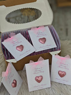 Box of pink heart-shaped favors with personalized cards on a textured surface.