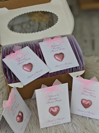 Box of pink heart-shaped favors with personalized cards on a textured surface.