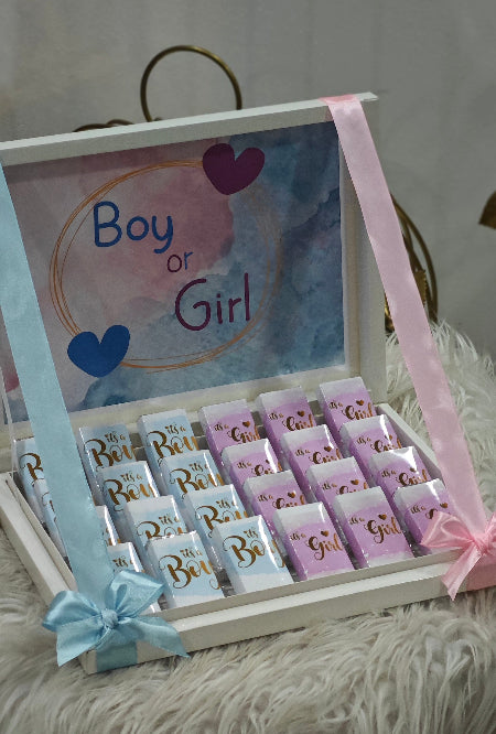Baby gender reveal box with chocolate bars and 'Boy or Girl' card on a fluffy surface.