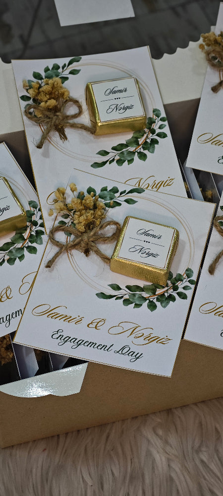 Engagement invitation cards with gold boxes and greenery on a textured surface