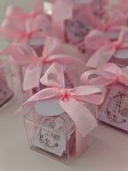 Small clear boxes with pink ribbons and decorative labels on a light background