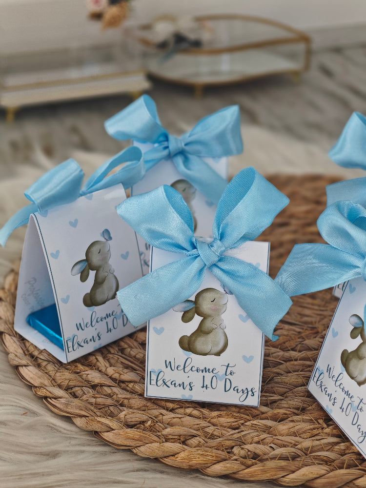 Small boxes with blue ribbons and baby shower-themed illustrations on a woven mat.