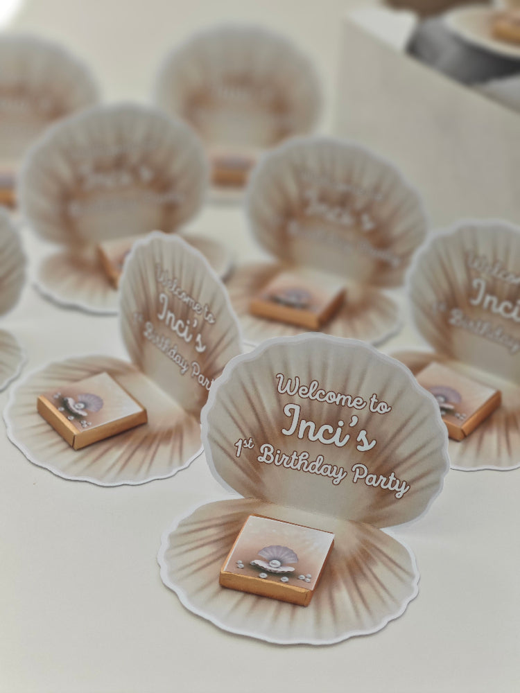 Seashell-shaped party favors with 'Welcome to Inci's 1st Birthday Party' text on a white surface.