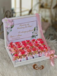 Decorative box with pink and red flowers, ribbons, and a card on a soft surface.