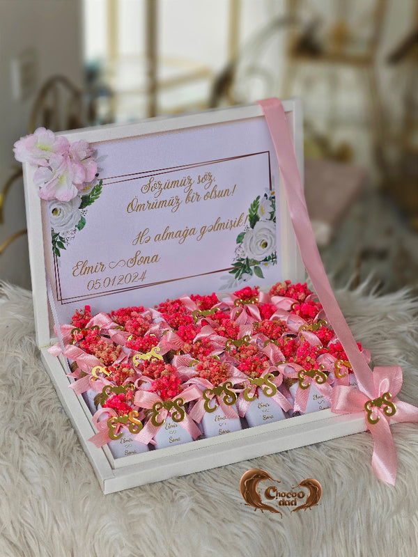 Decorative box with pink and red flowers, ribbons, and a card on a soft surface.