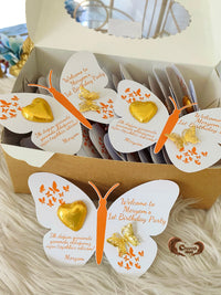 Butterfly-shaped party favors with gold hearts and orange antennae in a box.