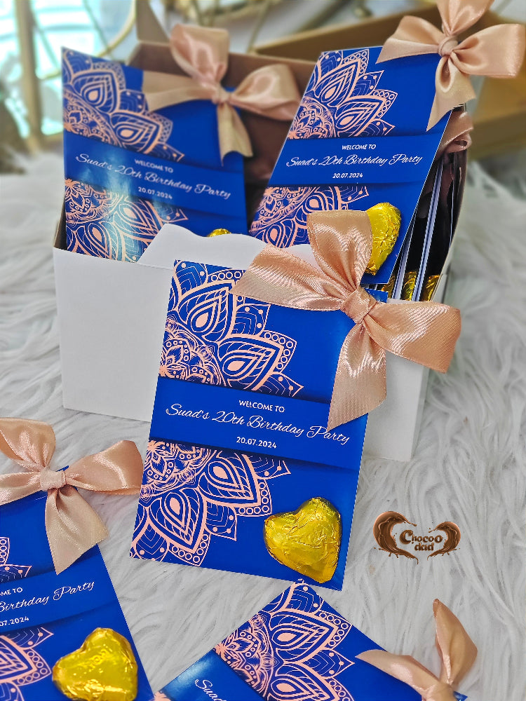 Blue birthday party invitations with pink ribbons and chocolate candies on a textured surface.