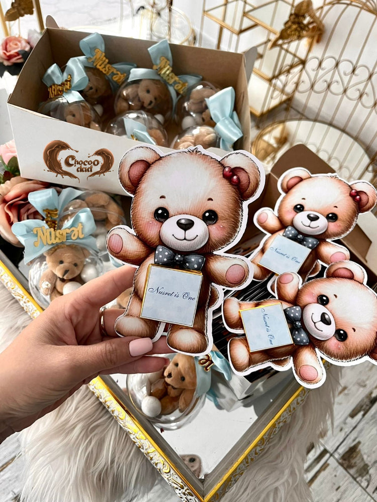 Sticker of a teddy bear with a bow tie held by a hand, with a box of similar stickers in the background.